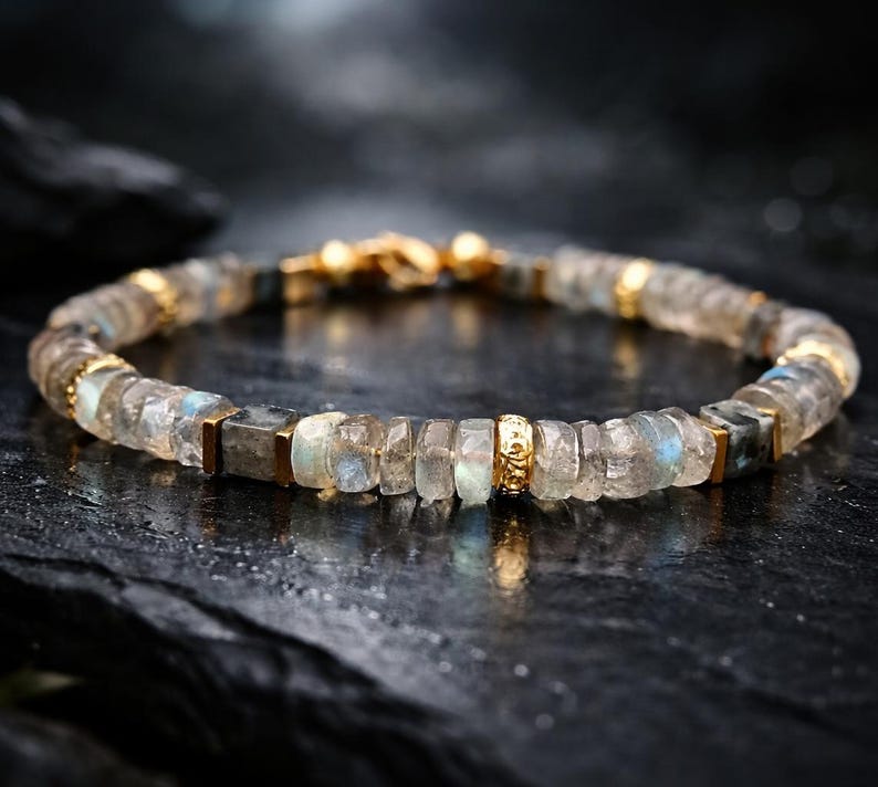 May include: A beaded bracelet featuring iridescent, rectangular and round-cut stones in shades of gray, blue, and clear. The bracelet is accented with gold-colored metal spacers and a clasp. The jewelry is displayed on a dark, textured surface.