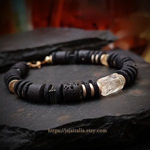 May include: A black lava stone bracelet with gold-colored accents and a clear crystal centerpiece. The bracelet is strung on a black cord and has a gold clasp. The bracelet is displayed on a dark, textured surface.