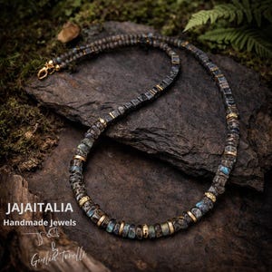 May include: A handcrafted necklace featuring dark gray and blue-green gemstone beads with gold-colored accents. The necklace is displayed on a dark stone surface. The text "JAJAITALIA Handmade Jewels" is visible.