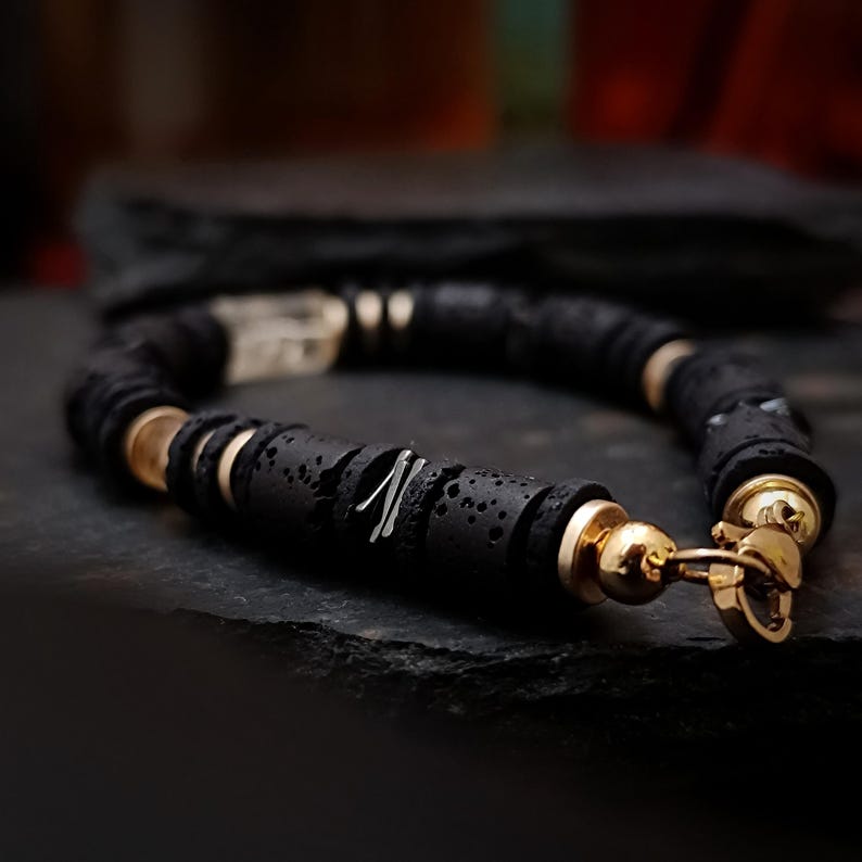 May include: Black lava stone bracelet with gold-colored accents. The bracelet features round, textured black beads interspersed with gold-colored spacers and a gold clasp. A silver-colored accent is visible on one of the black beads.