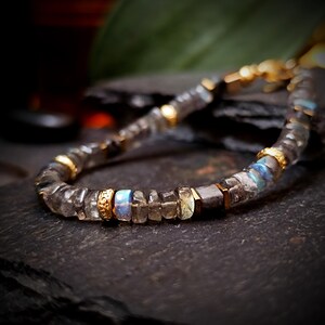 May include: A close-up of a beaded bracelet featuring gray and clear gemstone beads with gold-colored accents. The bracelet has a mix of rectangular and round beads, with some displaying blue and yellow iridescence. The bracelet is on a dark surface.