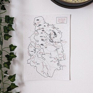 May include: A detailed map print of the Peak District, featuring hand-drawn illustrations of the region's towns, rivers, and landmarks. The map is in black and white with red accents, and the title "THE PEAK DISTRICT" is displayed at the top.