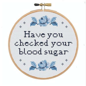 Può includere: Un telaio da ricamo a punto croce con la scritta "Hai controllato la tua glicemia?" in blu scuro. Il disegno include elementi floreali blu e segni più. Il telaio in legno è rotondo.