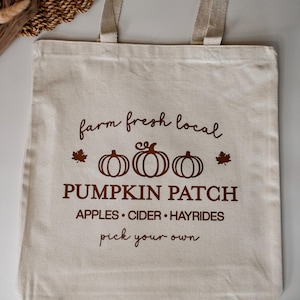 May include: A beige canvas tote bag with brown text that reads "farm fresh local" above three pumpkin illustrations and "PUMPKIN PATCH". Additional text includes "APPLES • CIDER • HAYRIDES" and "pick your own". The bag has beige handles.