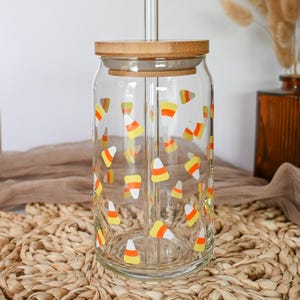 Könnte beinhalten: Ein durchsichtiger Glasbecher mit Bambusdeckel und einem durchsichtigen Glasstrohhalm. Der Becher ist mit einem Muster aus gelben, orangefarbenen und weißen Zuckermais verziert. Der Becher steht auf einer gewebten Matte.