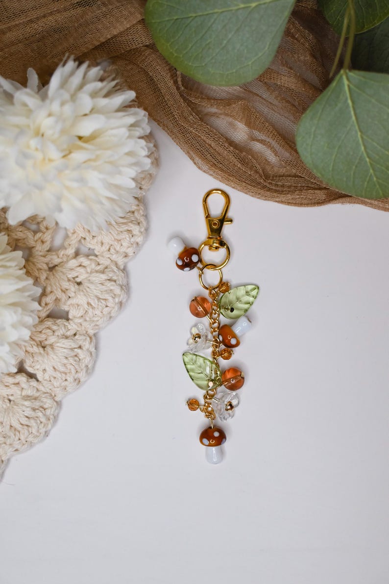 May include: A gold-colored keychain with a charm featuring mushroom and leaf-shaped beads. The charm includes orange, white, and clear beads, and green leaf-shaped beads. The keychain is on a white surface with a beige crocheted item and greenery in the background.