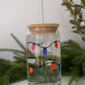 May include: A clear glass jar with a bamboo lid and a glass straw. The jar is filled with ice and water and has a design of colorful Christmas lights on the side.