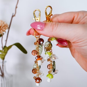 May include: Gold-colored keychain with dangling charms. The charms include mushroom, leaf, and bead designs in shades of brown, green, and clear. The keychain is held up by a hand with pink nail polish.