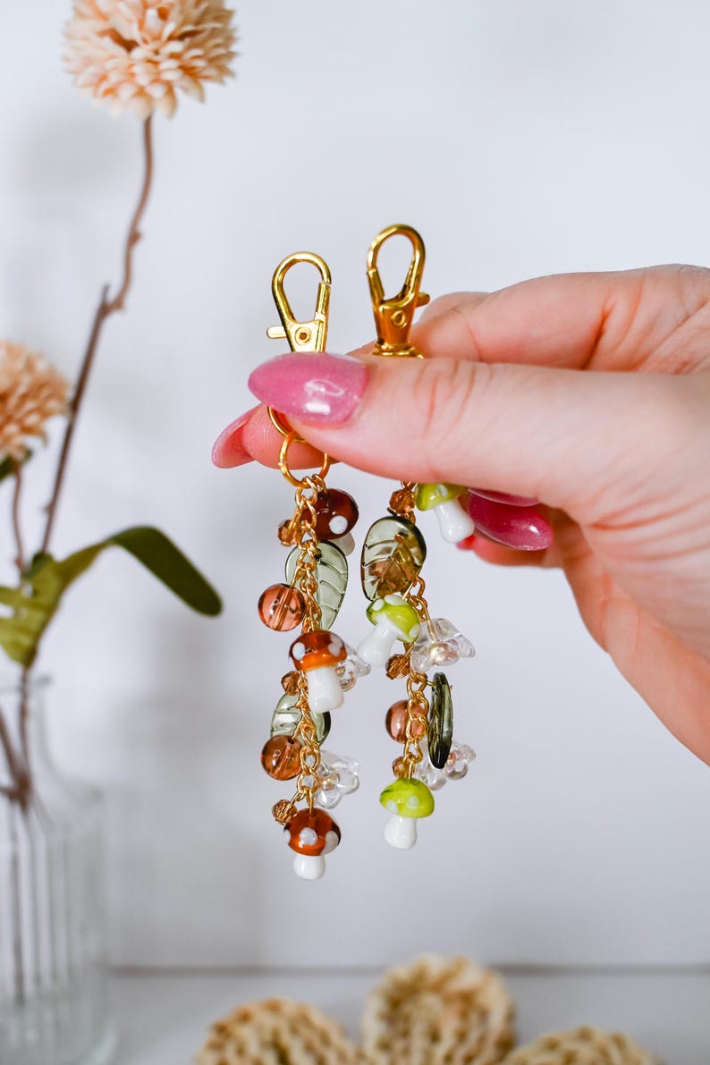May include: Gold-colored keychain with dangling charms. The charms include mushroom, leaf, and bead designs in shades of brown, green, and clear. The keychain is held up by a hand with pink nail polish.