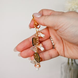 May include: A gold-colored keychain with a lobster clasp, adorned with mushroom charms, clear and amber-colored beads, and leaf-shaped charms. The keychain is held in a hand against a soft, neutral background.