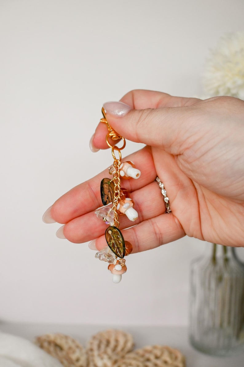 May include: A gold-colored keychain with a lobster clasp, adorned with mushroom charms, clear and amber-colored beads, and leaf-shaped charms. The keychain is held in a hand against a soft, neutral background.