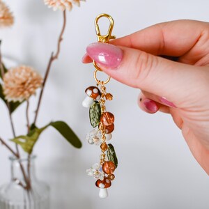 May include: A gold-colored keychain charm featuring mushroom, leaf, and bead accents. The charm includes small white and brown mushroom pendants, green leaf-shaped beads, and various orange and clear beads. The charm is held by a gold clasp.