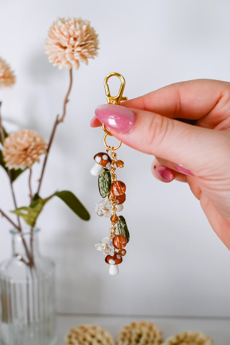 May include: A gold-colored keychain charm featuring mushroom, leaf, and bead accents. The charm includes small white and brown mushroom pendants, green leaf-shaped beads, and various orange and clear beads. The charm is held by a gold clasp.