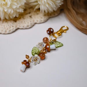 May include: A whimsical keychain featuring mushroom, leaf, and flower charms. The charms are in shades of brown, white, green, and clear, with gold-colored accents. The keychain has a gold-colored clasp.