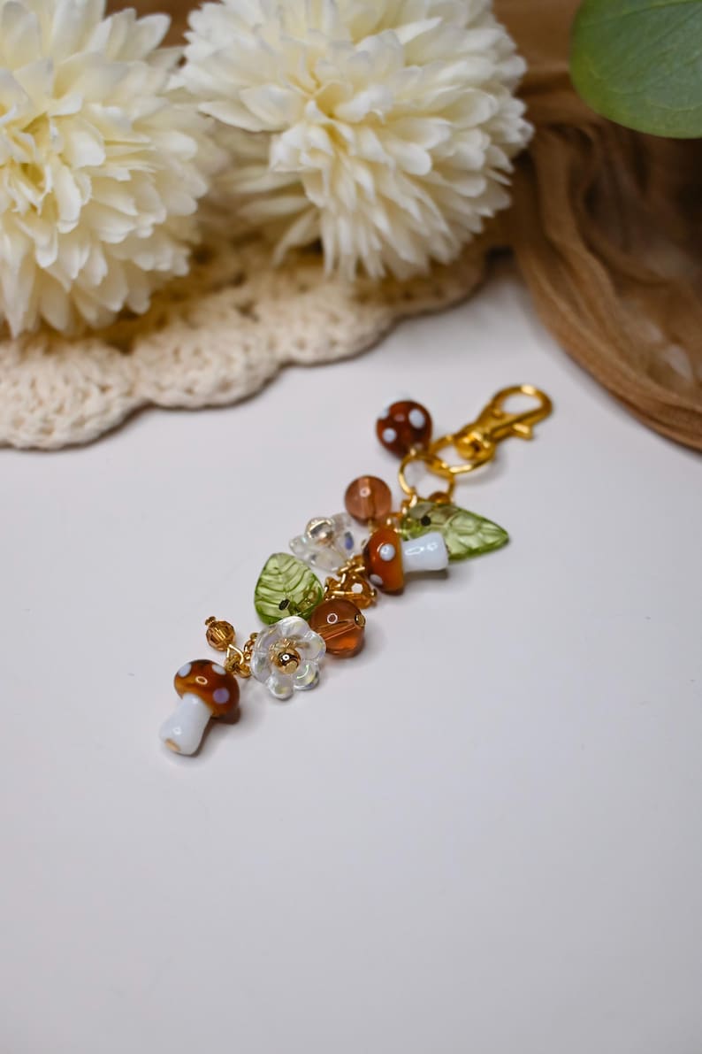 May include: A whimsical keychain featuring mushroom, leaf, and flower charms. The charms are in shades of brown, white, green, and clear, with gold-colored accents. The keychain has a gold-colored clasp.
