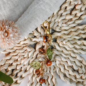 May include: A gold-colored keychain with a charm featuring mushroom and leaf-shaped beads. The charm includes orange, brown, and clear beads, along with green leaf accents. The keychain is set against a woven, light-colored background.