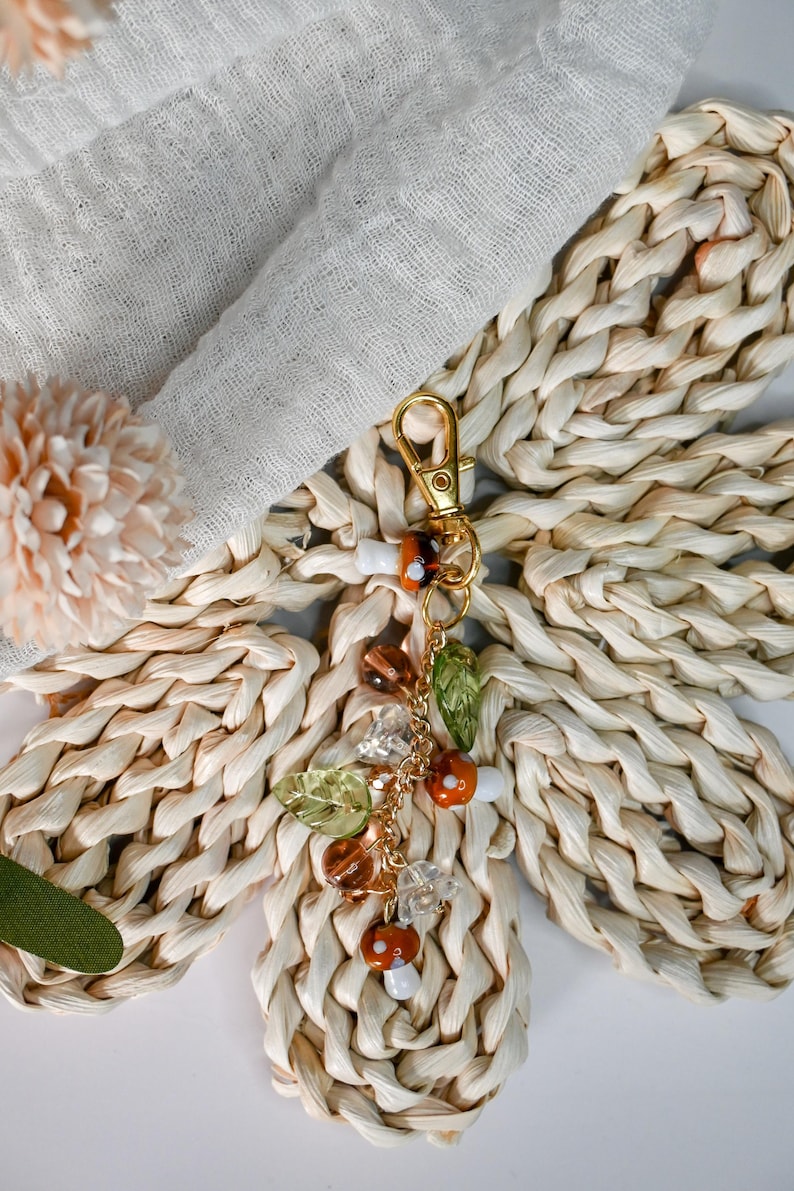 May include: A gold-colored keychain with a charm featuring mushroom and leaf-shaped beads. The charm includes orange, brown, and clear beads, along with green leaf accents. The keychain is set against a woven, light-colored background.