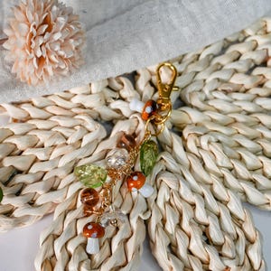 May include: A gold-colored keychain with mushroom and leaf charms. The charms include orange and white mushrooms, green leaves, and clear and brown beads. The keychain is attached to a gold-colored clasp.