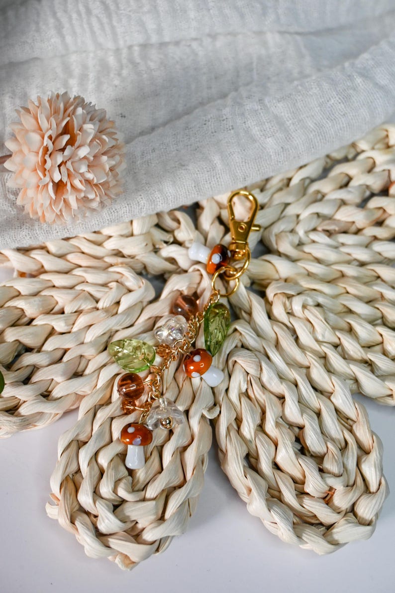 May include: A gold-colored keychain with mushroom and leaf charms. The charms include orange and white mushrooms, green leaves, and clear and brown beads. The keychain is attached to a gold-colored clasp.
