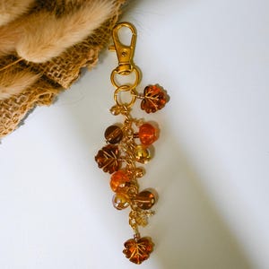 May include: A gold-toned metal keychain featuring a lobster clasp and a dangling cluster of amber, orange, and brown beads. The charms include translucent leaf-shaped beads and round spherical beads attached to a central gold-colored chain.