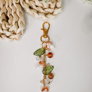 May include: A gold-colored keychain with a lobster clasp, adorned with mushroom charms, green leaf beads, and amber-colored beads. The keychain is suspended on a gold chain, creating a whimsical and nature-inspired accessory.