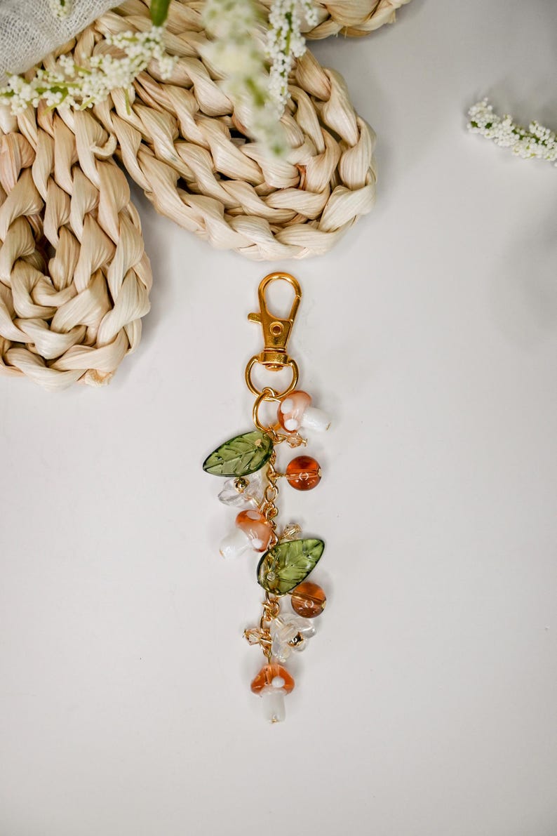 May include: A gold-colored keychain with a lobster clasp, adorned with mushroom charms, green leaf beads, and amber-colored beads. The keychain is suspended on a gold chain, creating a whimsical and nature-inspired accessory.