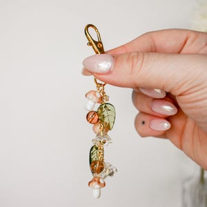 May include: A gold-colored keychain with a lobster clasp, adorned with decorative charms. The charms include mushroom, leaf, and crystal-like beads in shades of orange, green, and clear. The keychain is held in a person's hand.