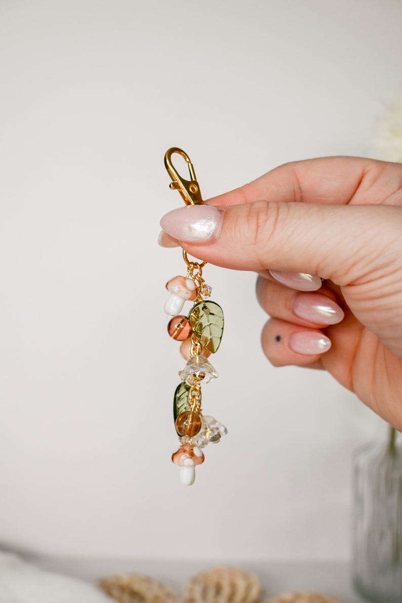 May include: A gold-colored keychain with a lobster clasp, adorned with decorative charms. The charms include mushroom, leaf, and crystal-like beads in shades of orange, green, and clear. The keychain is held in a person's hand.