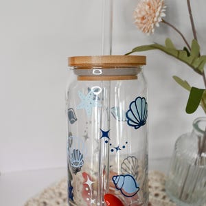 May include: A clear glass tumbler with a bamboo lid and a glass straw. The tumbler is decorated with blue and white seashell and starfish designs. The tumbler is held by a person with red nail polish.
