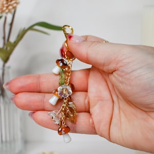May include: A gold-colored keychain with mushroom and floral charms. The charms include small, detailed mushrooms with brown and white caps, clear flower-shaped beads, and amber-colored leaf charms. The keychain is held in a hand.