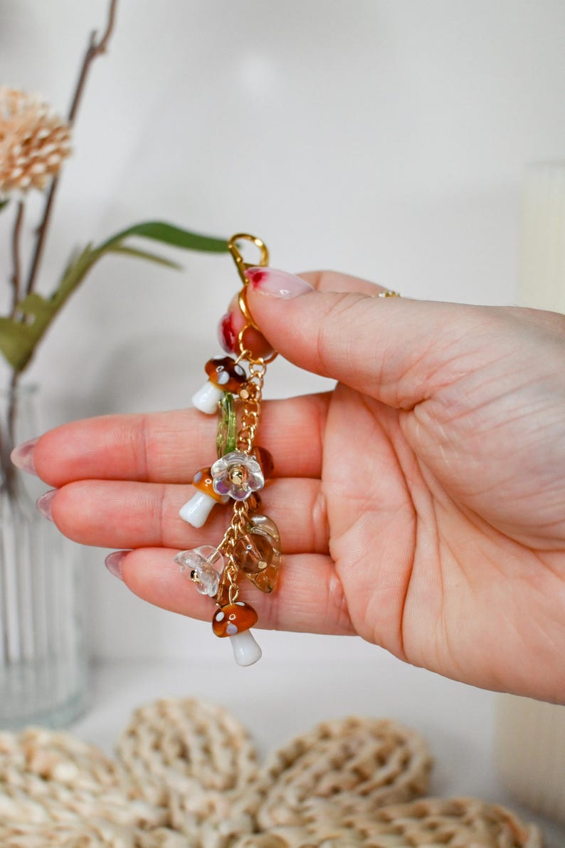 May include: A gold-colored keychain with mushroom and floral charms. The charms include small, detailed mushrooms with brown and white caps, clear flower-shaped beads, and amber-colored leaf charms. The keychain is held in a hand.