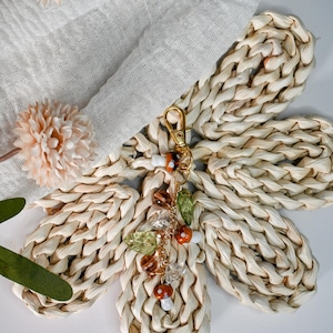 May include: A woven raffia flower with a gold-colored keychain. The keychain has a charm with mushroom and leaf accents. The flower is made of woven raffia and is beige in color. The charm has brown, orange, and clear beads.