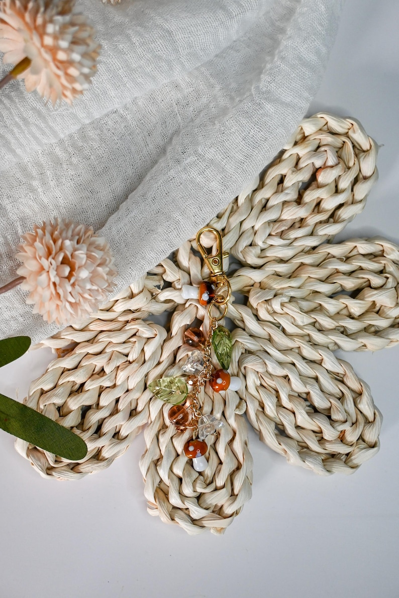 May include: A woven raffia flower with a gold-colored keychain. The keychain has a charm with mushroom and leaf accents. The flower is made of woven raffia and is beige in color. The charm has brown, orange, and clear beads.