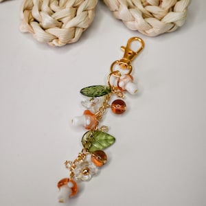 May include: A whimsical keychain featuring mushroom and leaf charms, along with orange and clear beads. The charms are strung on a gold-colored chain, and the keychain has a gold-colored clasp.