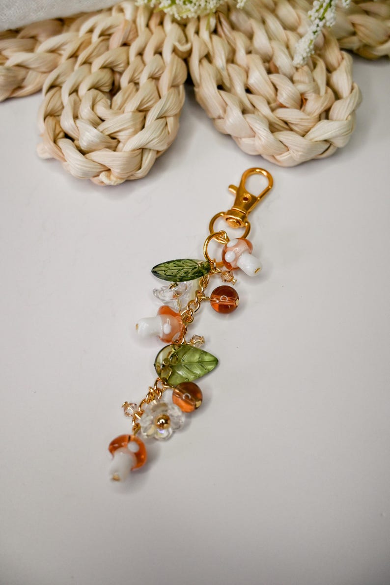 May include: A whimsical keychain featuring mushroom and leaf charms, along with orange and clear beads. The charms are strung on a gold-colored chain, and the keychain has a gold-colored clasp.