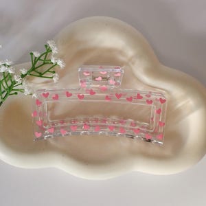 Puede incluir: Una pinza para el pelo de plástico transparente con un patrón de corazones rosas. La pinza está diseñada para sujetar el pelo.