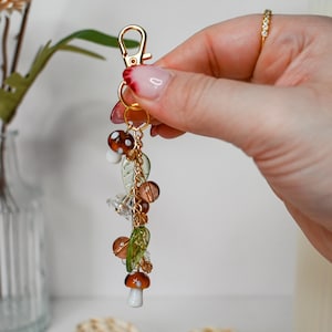 May include: A gold-colored keychain with a lobster clasp, adorned with mushroom and leaf charms. The charms are made of glass and beads in shades of brown, green, and clear. The keychain is a decorative accessory.