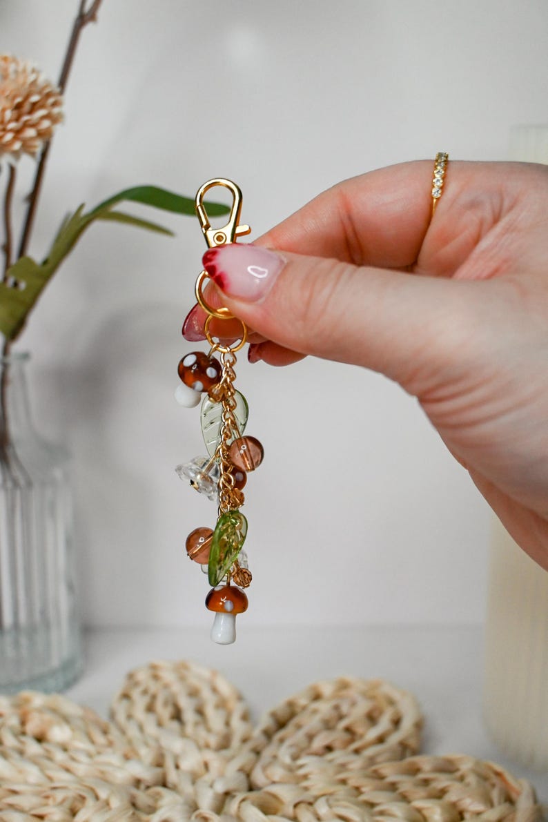 May include: A gold-colored keychain with a lobster clasp, adorned with mushroom and leaf charms. The charms are made of glass and beads in shades of brown, green, and clear. The keychain is a decorative accessory.