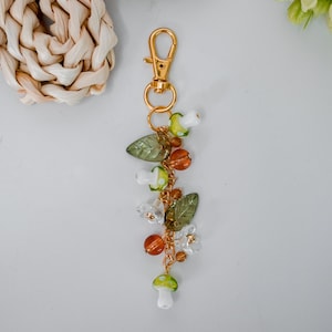 May include: A gold-colored keychain with a lobster clasp, adorned with a charm of glass beads, including green leaves, orange and clear beads, and white and green mushroom-shaped beads. The keychain is designed with a nature-inspired theme.