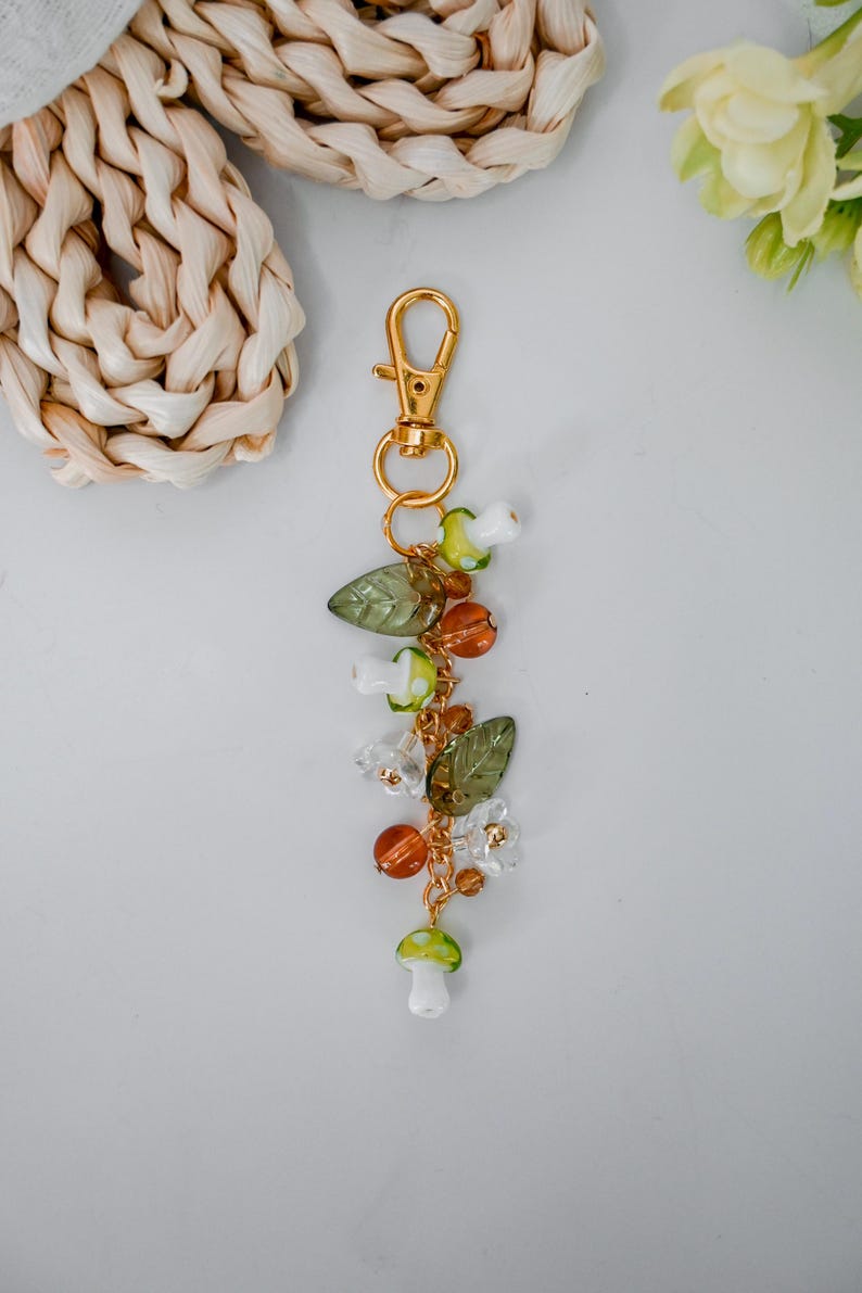 May include: A gold-colored keychain with a lobster clasp, adorned with a charm of glass beads, including green leaves, orange and clear beads, and white and green mushroom-shaped beads. The keychain is designed with a nature-inspired theme.
