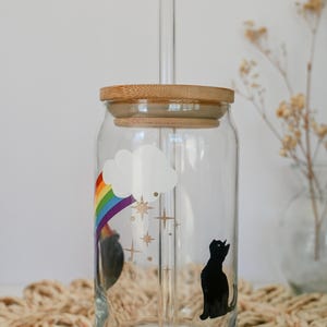 May include: A clear glass reusable water bottle with a rainbow, cloud, and stars design. The bottle has a bamboo lid and a glass straw. A black silhouette of a cat looking up at the stars is on the bottle.