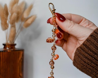 May include: A gold-toned metal keychain featuring a series of dangling beads in shades of amber, orange, and clear glass. The design includes small leaf-shaped charms and spherical beads strung together on a thin gold wire, ending with a lobster clasp.