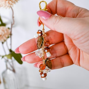 May include: A gold-colored keychain with a lobster clasp, adorned with mushroom and leaf charms. The charms are brown, white, and clear, with orange and gold beads. The keychain is held in a hand with pink nail polish.