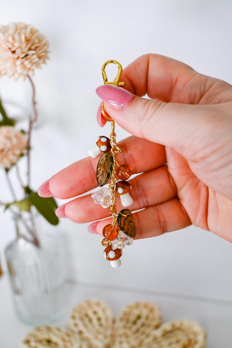 May include: A gold-colored keychain with a lobster clasp, adorned with mushroom and leaf charms. The charms are brown, white, and clear, with orange and gold beads. The keychain is held in a hand with pink nail polish.