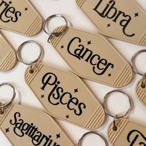 May include: A set of twelve beige keychains with silver key rings. Each keychain features a different zodiac sign in black lettering, including Leo, Libra, Cancer, Pisces, Sagittarius, Taurus, and Aquarius.