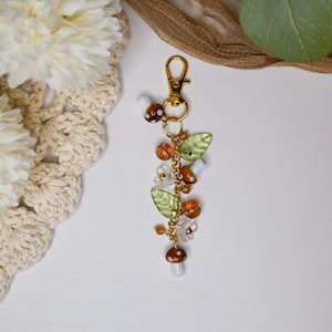 May include: A gold-colored keychain with a charm featuring mushroom and leaf accents. The charm includes brown and white mushroom beads, green leaf beads, and orange and clear crystal beads. The keychain is on a white surface.