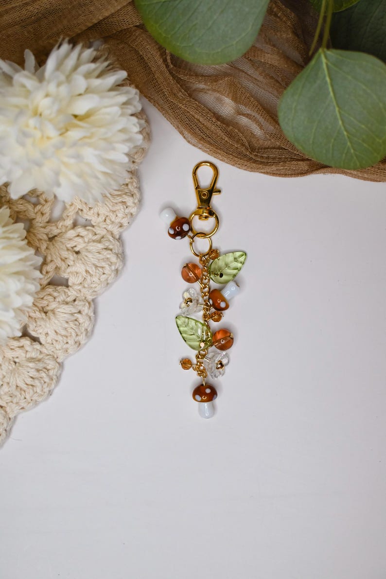 May include: A gold-colored keychain with a charm featuring mushroom and leaf accents. The charm includes brown and white mushroom beads, green leaf beads, and orange and clear crystal beads. The keychain is on a white surface.