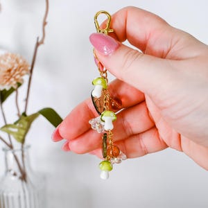 May include: A gold-colored keychain with a lobster clasp, adorned with mushroom charms in white and green, along with clear and amber-colored beads. The keychain is held against a soft background, showcasing its whimsical design.