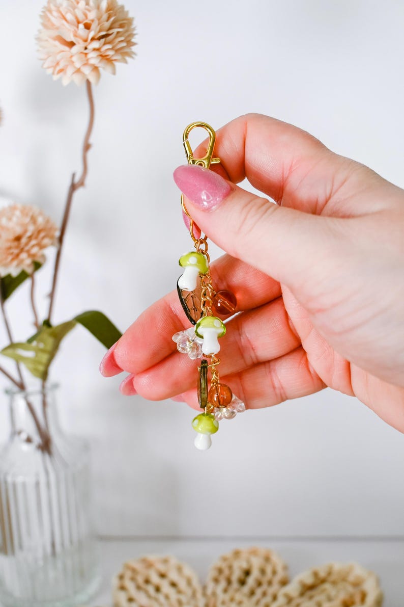 May include: A gold-colored keychain with a lobster clasp, adorned with mushroom charms in white and green, along with clear and amber-colored beads. The keychain is held against a soft background, showcasing its whimsical design.