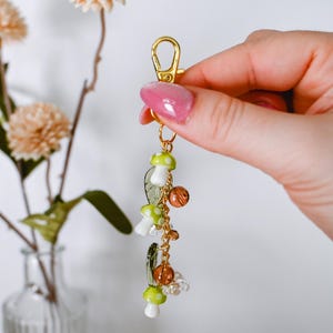 May include: A gold-colored keychain with a lobster clasp, adorned with mushroom charms in white and green, along with clear and amber-colored beads. The keychain is held in a hand, with a background of flowers in a vase.